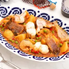 Caldeirada gallega servida en plato blanco con borde azul. Contiene trozos de pescado, patatas, guisantes y pimientos en una salsa rojiza.