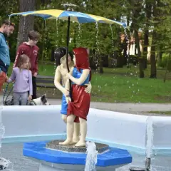 Rodzina spaceruje po ciechocinek deptak, podziwiając rzeźbę pary pod parasolem, z której tryska woda.