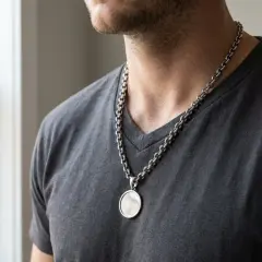 Mężczyzna w szarym t-shircie nosi masywny srebrny naszyjnik z okrągłą zawieszką. Łańcuszek typu rolo dodaje mu charakteru.
