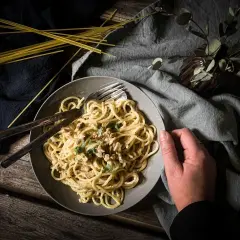 Teller mit cremiger, veganer Spaghetti Carbonara, garniert mit Kräutern. Daneben liegen ungekochte Spaghetti.
