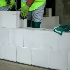 Budowa ściany z białych pustaków silikatowych. Ręce w zielonych rękawicach układają kolejny blok.