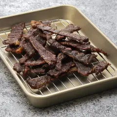 Stos cienkich, ciemnych plastrów wołowiny na metalowej kratce, gotowych do spożycia. Widać przyprawy, które nadają im smak, jak w klasycznej beef jerky marynata.