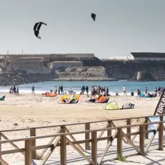 Plaża pełna kolorowych latawców i ludzi, gotowych do **kitesurfingu w Polsce**. W tle widać zabytkowe budowle.