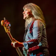 Suzi Quatro obecnie, z uśmiechem na twarzy, gra na gitarze basowej w dżinsowej kurtce.