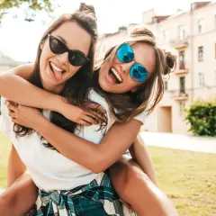 Dos amigas riendo y abrazándose, como si estuvieran escribiendo una carta a una amiga. Llevan gafas de sol y están al aire libre.