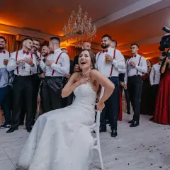 Radosna panna młoda na krześle, otoczona przez drużbów w czerwonych szelkach. Zastanawiasz się, ile kosztuje wesele na 50 osób?