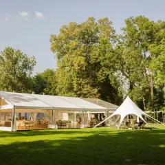 Eleganckie wesele w namiocie na trawie, wśród drzew. Wnętrze namiotu udekorowane stołami i krzesłami, gotowe na przyjęcie gości.