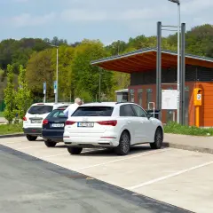 Stationnement sur une aire d'autoroute avec plusieurs voitures garées. Un homme est assis sur un banc rouge devant un bâtiment orange.