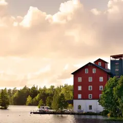 Skålleruds gård vid vattnet, med en röd byggnad och ett torn, omgiven av träd under en molnig himmel.