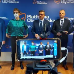 Pięciu mężczyzn w garniturach stoi przed niebieskim banerem z logo Platformy Obywatelskiej. Na pierwszym planie widać telefon nagrywający wydarzenie.