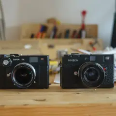 Two classic rangefinder cameras, a Leica CL and a Minolta, sit side-by-side on a wooden desk, ready for their next adventure.
