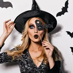 Kobieta w kapeluszu czarownicy z literami "Boo!" przed twarzą. Jej prosty makijaż czarownicy podkreśla oczy i usta.