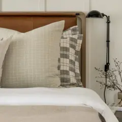 Studio McGee bed styling with layered pillows featuring subtle patterns and a plaid accent. A bedside lamp and delicate branches in a vase complete the serene look.