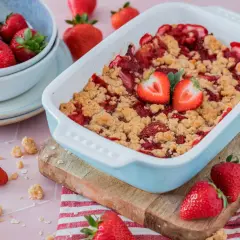 Ein frisch gebackener Erdbeer Crumble in einer hellblauen Auflaufform, garniert mit Erdbeerhälften. Daneben eine Schüssel mit Erdbeeren und verstreute Streusel.