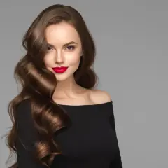 Mujer con labios rojos y espectaculares ondas pelo largo castaño, luciendo un vestido negro de hombros descubiertos.