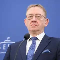 Mężczyzna w okularach i granatowym garniturze przed mikrofonem na tle niebieskiej ściany z logo Senatu.