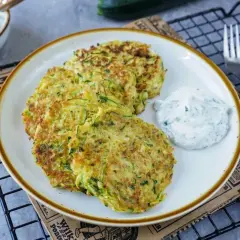 Drei goldbraune Zucchini-Pfannkuchen liegen auf einem Teller, daneben ein Klecks Kräuterquark. Ein leckeres Gericht!