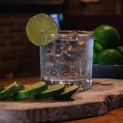 Erfrischender Gin Tonic Water mit Eis und Limettenscheiben auf einem Holzbrett. Mehrere Limetten liegen in einer Schale daneben.