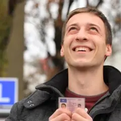 Szczęśliwy młody mężczyzna trzyma prawo jazdy, sprawdzając czy jest do odbioru. W tle niebieski znak "L".