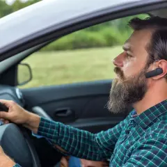 Mężczyzna z brodą i słuchawką Bluetooth w uchu prowadzi samochód. Dowiedz się, jak sparować telefon z samochodem, by bezpiecznie rozmawiać.