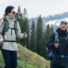 Para w odpowiednim ubraniu w góry, z kijkami trekkingowymi, cieszy się wędrówką pośród zielonych wzgórz i ośnieżonych szczytów.