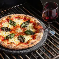 Pizza mit langer Gehzeit auf dem Grill, belegt mit Tomaten, Mozzarella und Basilikum, daneben ein Glas Rotwein.
