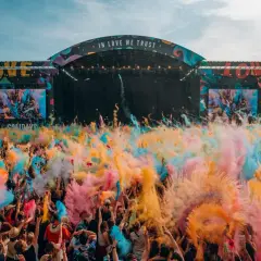 Une foule en liesse lance de la poudre colorée pour créer un festival de musique inoubliable. L'ambiance est électrique sous la scène "IN LOVE WE TRUST".