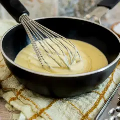 Eine cremige low carb bechamel wird in einer Pfanne mit einem Schneebesen verrührt. Daneben stehen Schüsseln mit Sahne, Salz und Pfefferkörnern.