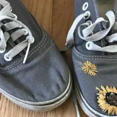 Paire de chaussures grises avec des lacets blancs. L'une des chaussures est ornée de broderies de tournesols, donnant un air bohème à ces chaussures trouées.