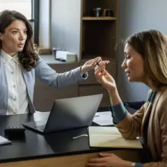 Dwie kobiety w biurze, jedna gestykuluje, druga słucha. Widać laptop, dokumenty i kubek. Atmosfera sugeruje stres w pracy.