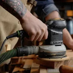 Main d'un artisan utilisant un outil pour poncer du bois. L'outil est relié à un aspirateur par un tuyau vert.