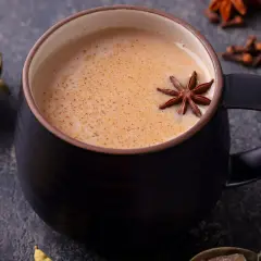Aromaticzny napój, idealny na chłodne dni. Gorący **masala chai** w czarnej filiżance, ozdobiony anyżem. Obok liście herbaty, kardamon i goździki.