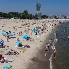Słoneczna plaża w jednej z miejscowości koło Gdyni, pełna wypoczywających ludzi. W tle widać drapacz chmur i żaglowce.