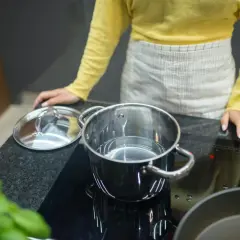 Kobieta w fartuchu i żółtej bluzce stoi przy płycie indukcyjnej, na której stoi garnek z wodą. Pokazuje, jak wyczyścić płytę indukcyjną.