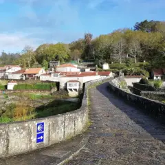 Antiguo puente de piedra con señal de peregrinación. Al fondo, un pueblo con casas de tejados rojos y vegetación exuberante. ¡Ponte Maceira que ver!