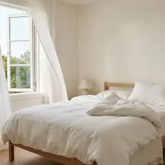Ein helles Schlafzimmer mit einem offenen Fenster und weißen Vorhängen. Ein Thermometer auf dem Nachttisch zeigt die ideale Temperatur, bei welcher Temperatur sollte man schlafen.