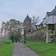 Ein Paar spaziert auf einem Weg entlang der alten Stadtmauer von Zons am Rhein. Ein Weidenbaum und ein Wehrturm prägen die Szenerie.