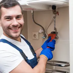 Uśmiechnięty hydraulik w niebieskim kombinezonie i rękawiczkach dokręca kluczem bojler. Zastanawiasz się, czy wyłączać bojler na wyjazd? On wie!