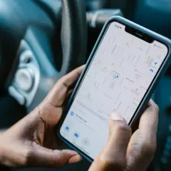 Dłonie trzymają telefon z mapą, pokazującą aplikację do udostępniania lokalizacji. Widać fragment kierownicy samochodu.