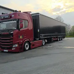 Czerwona Scania z naczepą gotowa do przewozu drobnicowego. Obok stoi niebieska toaleta przenośna.