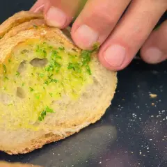 Un trozo de pan rústico con una generosa capa de mantequilla de ajo y perejil picado, listo para ser horneado.