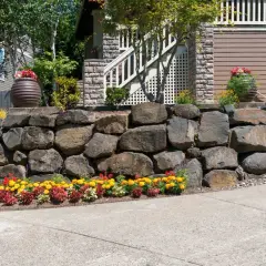 Natural stone retaining wall ideas: a sturdy wall of large rocks anchors a sloped yard, adorned with colorful flowers and planters.
