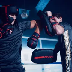 Two men in boxing gear practice sparring in a ring. One wears a headguard and gloves, throwing a punch at a pad held by the other.