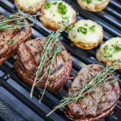 Saftige Rindersteaks mit Kräutern auf dem Holzkohlegrill. Daneben liegen gefüllte Champignons.