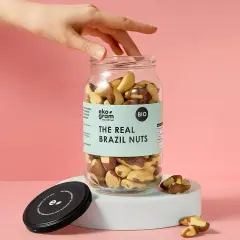 Dłoń sięga po słoik pełen orzechów brazylijskich. Na etykiecie widnieje napis "THE REAL BRAZIL NUTS".