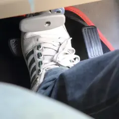 Un pied en basket Adidas appuie sur la pédale de frein, qui semble un peu molle. Le tapis de sol est noir avec un contour rouge.