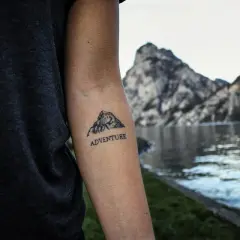Tatuaż góry z napisem "ADVENTURE" na ramieniu, na tle jeziora i skalistych szczytów.