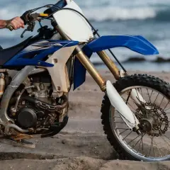 Persona sentada en una moto de cross azul y blanca en la arena, con el mar al fondo. ¿Cuánto paga un desguace por una moto?