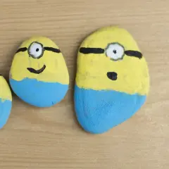 Trzy kamienie polne pomalowane na żółto i niebiesko, przypominające Minionki. Zabawne malowanie kamieni polnych.