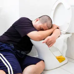 Mężczyzna śpi na muszli klozetowej, trzymając butelkę. Widać, że doświadczył wymiotów po alkoholu.